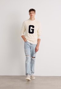 Cream sweater featuring a black "G" logo, paired with light blue, distressed jeans and beige sneakers. Simple, casual design.