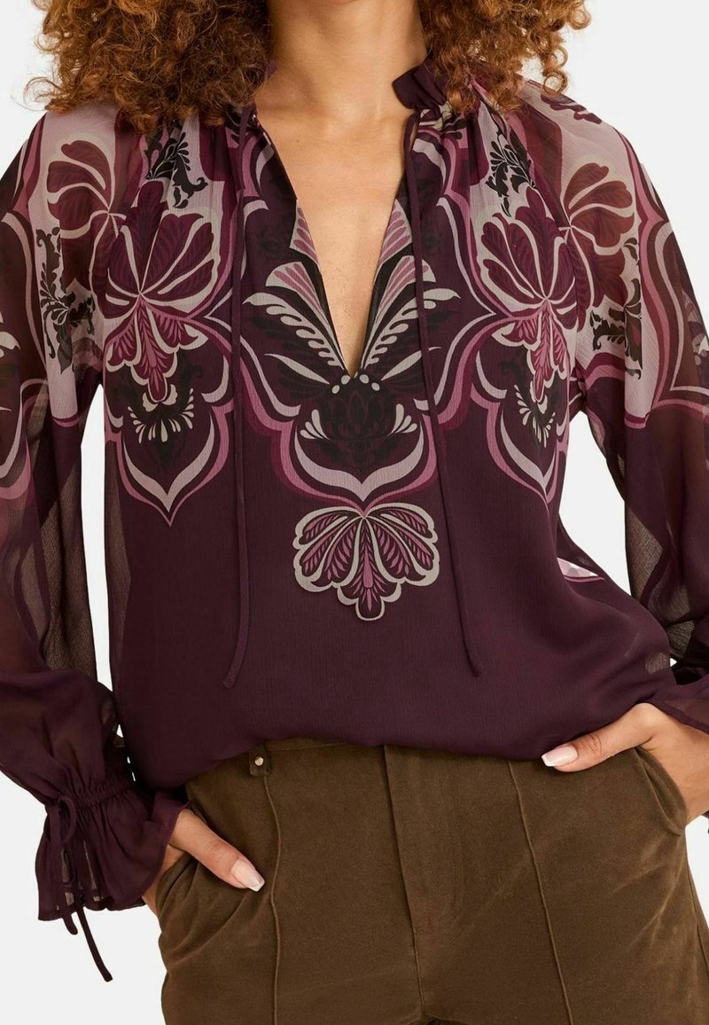 Femme portant un chemisier violet transparent à manches longues avec des motifs floraux et un pantalon vert olive, mains dans les poches, cheveux bouclés visibles.
