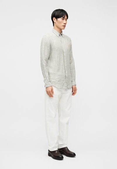 Άνδρας που  στέκεται φορώντας ένα ανοιχτό γκρι ριγωτό πουκάμισο με κουμπιά, λευκό παντελόνι και μαύρα δερμάτινα παπούτσια, μπροστά σε απλό λευκό φόντο.