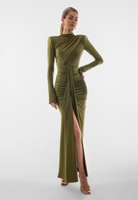Olivově zelené šaty s dlouhým rukávem, bočním rozparkem, sklady v pase a hladkým povrchem. Stylizováno na modelce na bílém pozadí.