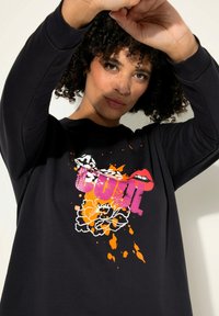 Maglietta nera a maniche lunghe con un design grafico colorato, con la parola "CULT" in rosa, labbra e motivi floreali arancioni e neri.