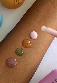 Fyra färgstarka droppar vätska: lila, grön, orange och rosa på en brun hudyta, med en rosa applikator som pekar på dem.