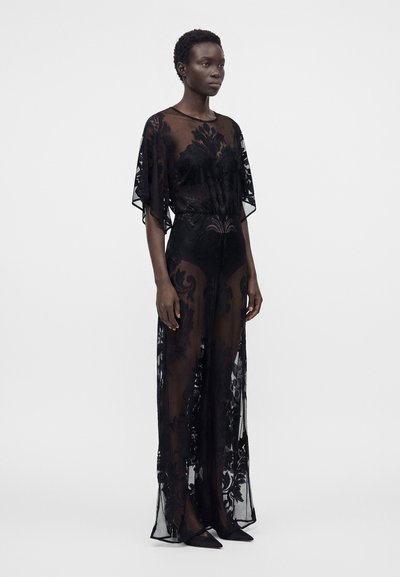Femme portant une robe noire transparente à motifs floraux, debout les bras détendus, devant un fond clair uni.