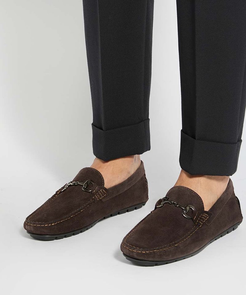Mocassins en daim marron avec détail métallique portés avec un pantalon noir retroussé et sans chaussettes visibles sur un sol gris clair.