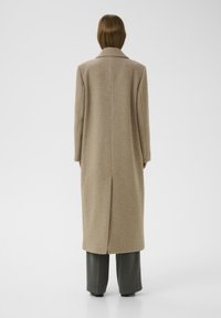 Abrigo de lana beige con corte recto, mangas largas y una abertura en la parte trasera. Presenta un cuello clásico y cae a mitad de la pantorrilla, combinado con pantalones oscuros.