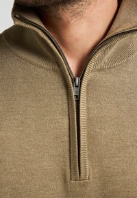 Beige Kapuzenpullover mit Reißverschluss, der eine Strickstruktur und einen Metallreißverschluss aufweist. Nahaufnahme mit Fokus auf den Reißverschluss und die Stoffdetails.