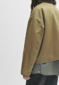 Veste en coton vert olive à coupe décontractée, avec un col en velours côtelé marron, des boutons pression et une chemise noire et blanche à motif en dessous.