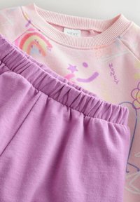 Jasnoróżowa bluza z pastelowymi grafikami, w komplecie z szarymi spodniami dresowymi na elastycznej talii w kolorze lawendowym. Wykonana z miękkiej bawełny.