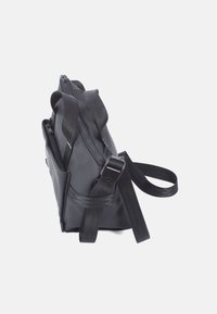 Borsa nera a più tasche realizzata in materiale morbido, dotata di chiusura con zip, spallacci regolabili e una forma strutturata con dettagli del logo.