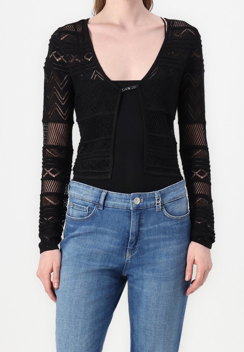 Czarny dziergany krótki kardigan z długimi rękawami, zdobiony geometrycznymi wzorami i głębokim dekoltem w kształcie litery V. Połączony z jasnoniebieskimi dżinsami.