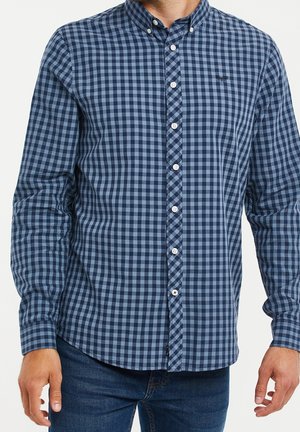 Mann mit blauem und marineblau kariertem Hemd mit Knöpfen und umklappbarem Kragen sowie dunkelblauer Jeans, steht mit entspannten Armen.