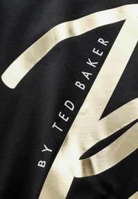 Černá látka s výrazným zlatým fóliovým písmem a designem, s textem "BY TED BAKER" na výrazně hladkém povrchu.