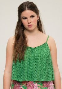 Grön spetskort topp med tunna band, med en mönstrad textur. Den nedre kanten har en rak skärning, och livliga färger syns nedanför.