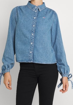 Mujer con una camisa de mezclilla azul con botones y mangas abullonadas atadas en las muñecas, combinada con pantalones negros.