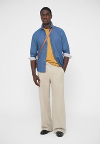 Kék farmering sárga póló felett, bézs színű, bő szárú nadrággal és fekete cipővel párosítva. Egy barna oldaltáska pántja is látható.