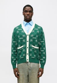 Cardigan verde feito de tecido de malha, apresentando um acabamento creme, bolsos e um design padrão com figuras de cartoon e texto em branco.