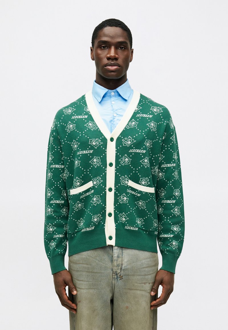 Cardigan verde feito de tecido de malha, apresentando um acabamento creme, bolsos e um design padrão com figuras de cartoon e texto em branco.