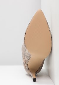 Spetsig tå högklackad sko gjord av texturerat, beige och brunt ormtrycksmaterial, med en slät, gräddfärgad sula och en smal svart klack.