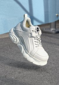 White sneakers with smooth leather upper, rounded thick rubber sole, and textured heels. Features lacing system and black branding accent.
