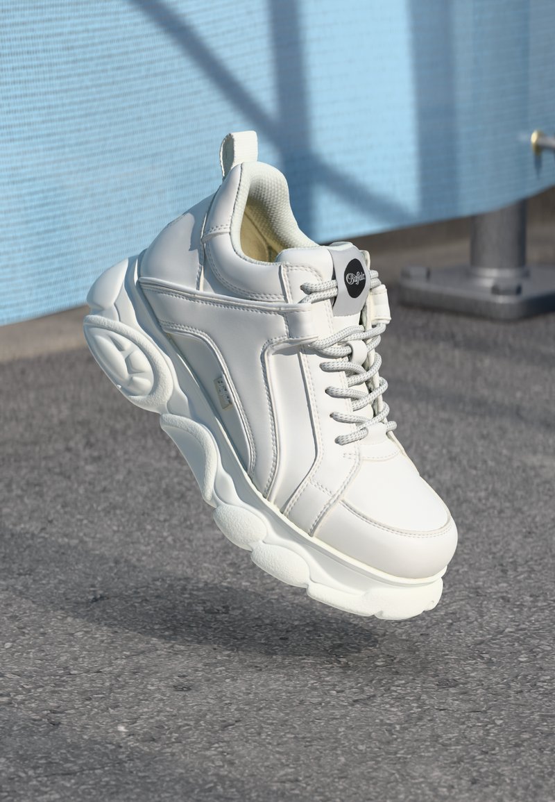 White sneakers with smooth leather upper, rounded thick rubber sole, and textured heels. Features lacing system and black branding accent.