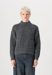 Grijze gebreide trui met een hoge kraag, twee voorzakken en een gestructureerde afwerking. Gecombineerd met losse denim broek.