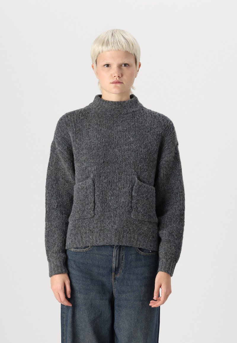 Grijze gebreide trui met een hoge kraag, twee voorzakken en een gestructureerde afwerking. Gecombineerd met losse denim broek.