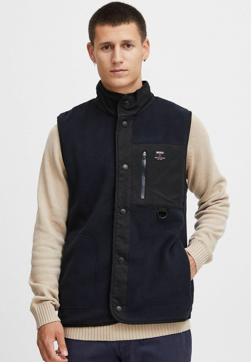 Chaleco de forro polar azul marino con una parte delantera de nailon negro, botones a presión, un bolsillo en el pecho con cremallera y dos bolsillos laterales; modelo de pie con suéter beige debajo.