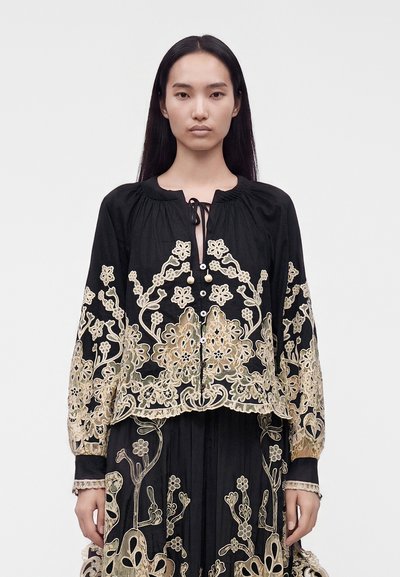 Femme portant un chemisier noir à manches longues avec broderie florale beige et une jupe assortie, debout devant un fond clair uni.