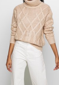 Beiger Strickpullover mit hohem Rollkragen, versehen mit einem Zopfmuster und gerippten Bündchen, kombiniert mit weißen weit geschnittenen Hosen.