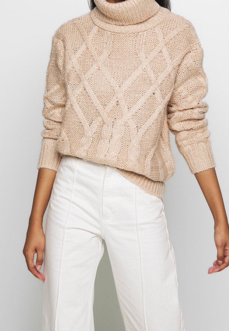 Beiger Strickpullover mit hohem Rollkragen, versehen mit einem Zopfmuster und gerippten Bündchen, kombiniert mit weißen weit geschnittenen Hosen.