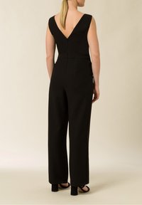 Svart jumpsuit med en figurnära midja, vida ben och djup V-design på ryggen. Slät textur med dragkedja bak och inga ytterligare utsmyckningar.