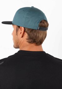Casquette turquoise avec un bord noir, fermeture snapback ajustable et tissu texturé. Portée par une personne en shirt noir, vue de derrière.