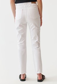 Vaqueros de mezclilla blanca con pierna recta, diseño de cinco bolsillos y pretina alta. Presenta un parche de cuero en la parte posterior. Se usan con sandalias negras.