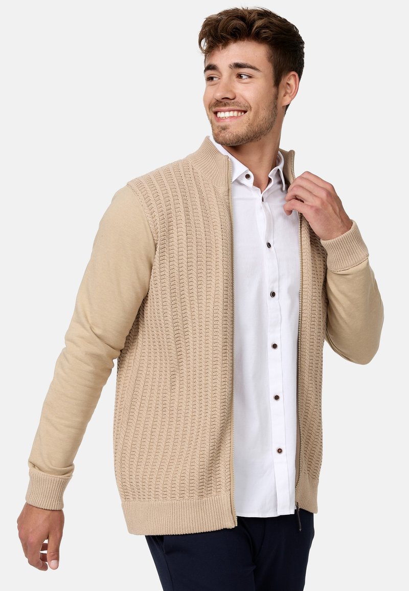Cardigan en tricot beige avec une texture côtelée, fermeture éclair et manches longues. Associé à une chemise blanche à boutons en dessous.