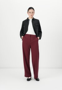 Woman standing with hands in pockets wearing black jacket, white top, burgundy wide-leg pants, black flats, and glasses against white background.