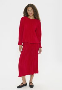 Donna con capelli ricci che indossa una maglia rossa a maniche lunghe con pieghe e una gonna midi abbinata, con scarpe flats nere, in piedi davanti a uno sfondo bianco.
