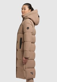 Long manteau en duvet beige avec une capuche, présentant des sections matelassées, une poche zippée noire et un patch de logo sur la manche. Texture lisse et isolée.