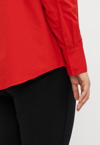 Camicia rossa con bottoni, a maniche lunghe, dal tessuto morbido e con orlo arrotondato. Dotata di polsini con due bottoni per una chiusura sicura.