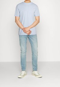 Mann trägt ein hellblaues T-Shirt mit kurzen Ärmeln, hellblaue eng anliegende Jeans und weiße High-Top-Sneaker und steht vor einem einfarbigen Hintergrund.