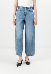 Mujer vistiendo jeans de pierna ancha de talle alto en azul claro y tacones slingback de punta negra, de pie contra un fondo blanco liso.