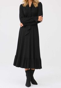 Femme portant une robe midi noire à manches longues avec un ourlet à volants et des bottines chaussettes noires assorties, debout les bras croisés.