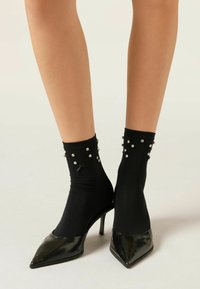 Czarne skarpetki do kostek z perłowymi akcentami i małymi kokardkami, w połączeniu z błyszczącymi czarnymi szpilkami. Gładkie wykończenie materiału, dopasowany krój.