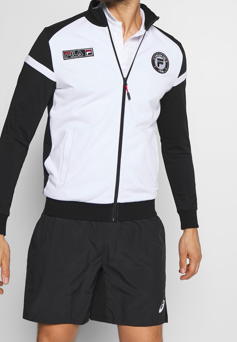 Giacca zip-up bianca e nera con polsini e vita a costine, caratterizzata da logo Fila ricamato e tasche laterali. Indossata con pantaloni corti neri.