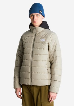 Junger Mann trägt eine beige Daunenjacke, blaue Mütze und olivgrüne Hose, steht vor einem einfarbigen hellgrauen Hintergrund.