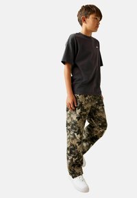 Tricou negru cu mânecă scurtă, asortat cu pantaloni largi cu model de camuflaj în nuanțe de verde și gri. Adidași albi completează ținuta.