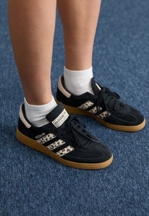 Jambes portant des chaussettes blanches montantes et des baskets noires Adidas Spezial avec des bandes imprimées léopard, debout sur un tapis bleu.