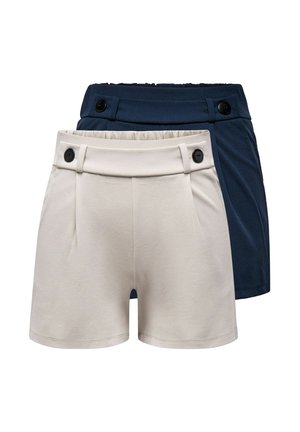 Frau in weißem ärmellosem Oberteil und dunkelblauen Shorts, die eine große schwarze Handtasche hält und schwarze offene Schuhe trägt, vor einfachem Hintergrund stehend.
