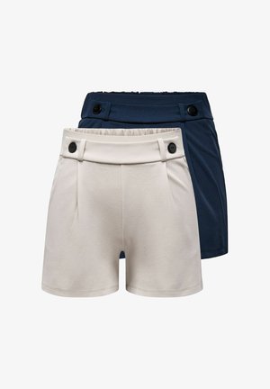 Frau in weißem ärmellosem Oberteil und dunkelblauen Shorts, die eine große schwarze Handtasche hält und schwarze offene Schuhe trägt, vor einfachem Hintergrund stehend.