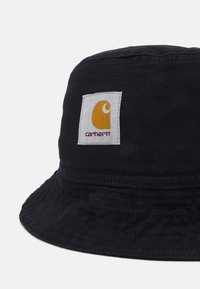 Chapeau seau noir en tissu texturé, avec un patch carré blanc portant un logo jaune et "carhartt" en texte foncé.