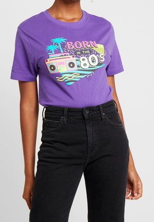 Lila T-Shirt mit buntem 80er-Jahre-Motiv, das einen Boombox, Palmen und den Text "Born in the 80's" zeigt, getragen mit schwarzen hoch taillierten Jeans.